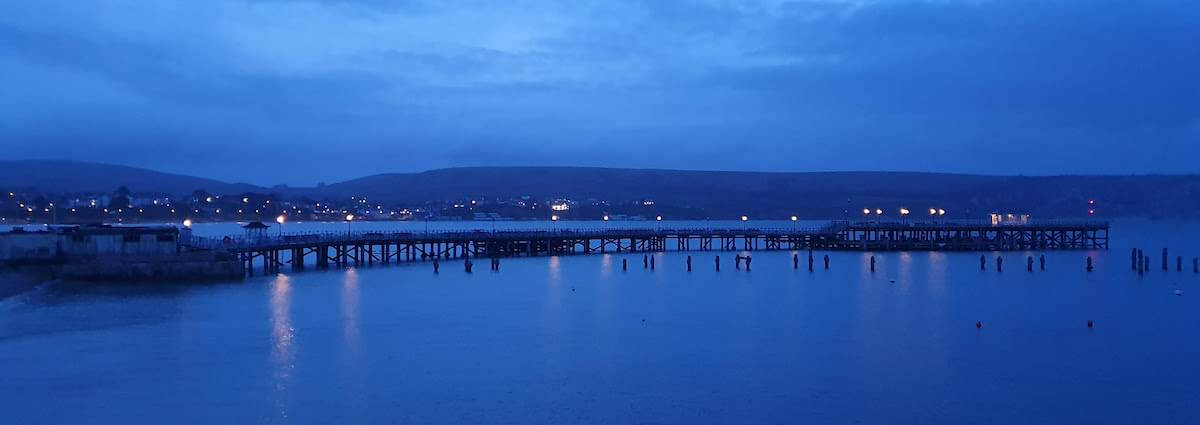 swanage pier at dusk with light reflecting into the calm waters in the foreground, and hills in the background