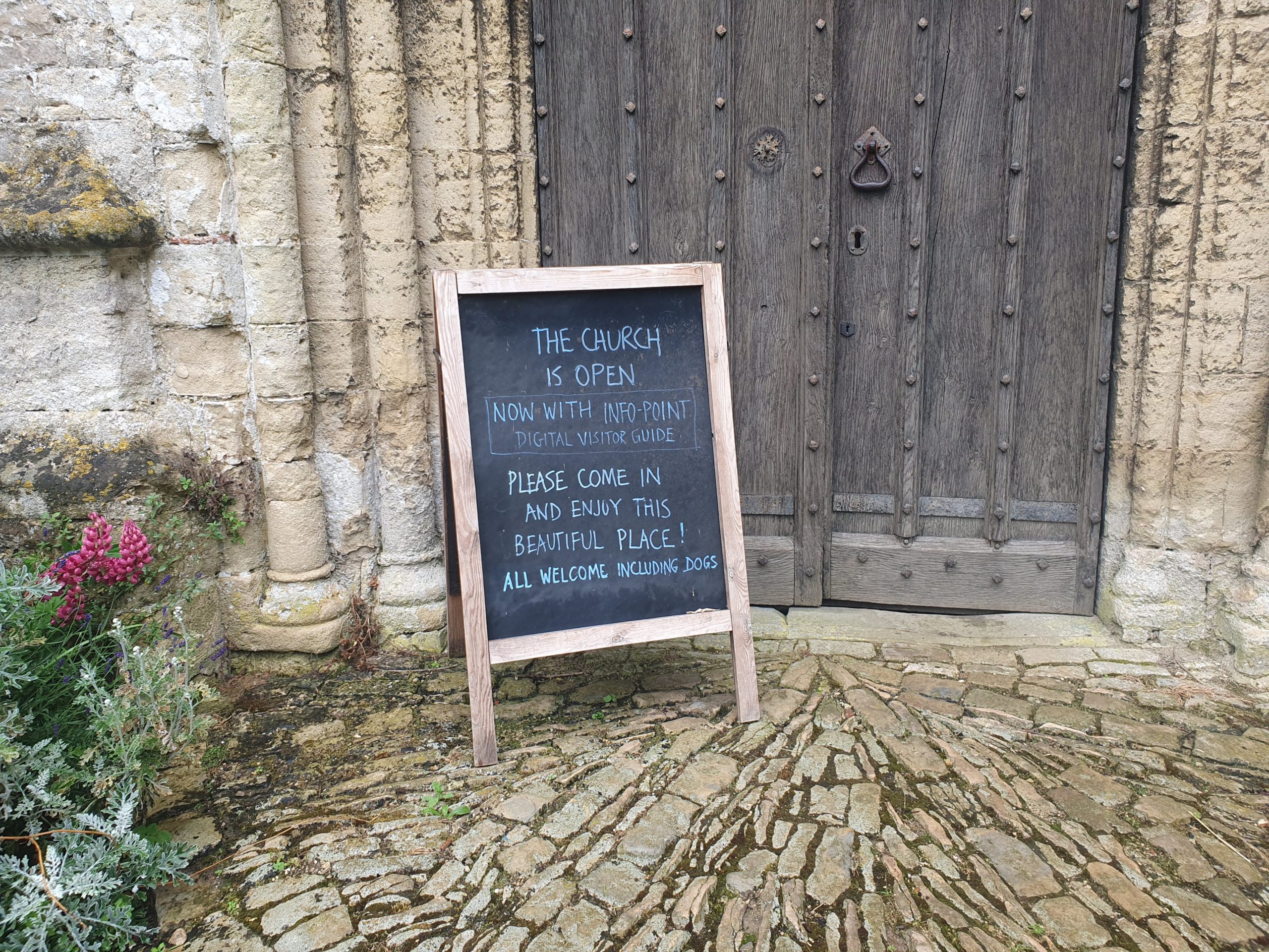 A chalk board with the words, "The church is open. Now with Info-Point digital visitor guide" stands on the cobbled pathway outside the closed wooden doorway of the church.