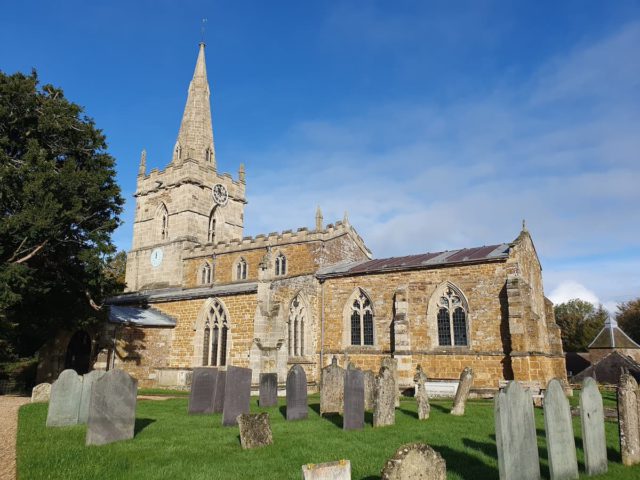 An external view of St John's church fills the image. A large tree on the left hugs against the beige spire of the tower. There is a blue sky. Gravestones can be seen amongst the grass in the foreground